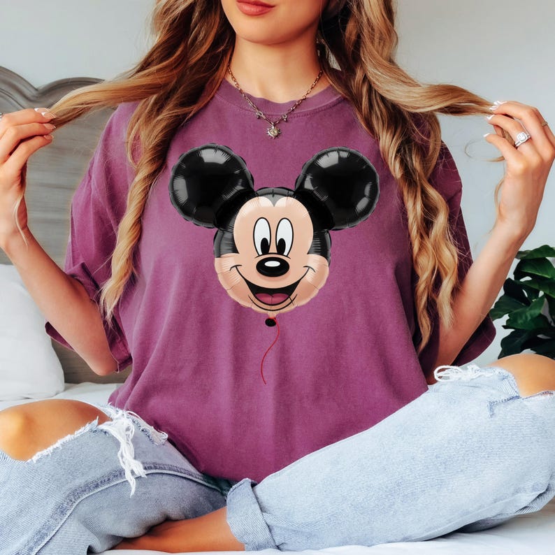 Puede incluir: Camiseta granate con un dise&ntilde;o de globo de Mickey Mouse. El globo tiene orejas negras, una cara color canela y una expresi&oacute;n sonriente. Una cuerda roja cuelga de la parte inferior del globo.