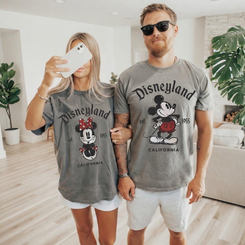 May include: Two gray t-shirts with the Disneyland logo and Mickey and Minnie Mouse graphics. The shirts have the text "Disneyland est. 1955 California". The people are wearing white shorts.