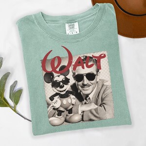 Puede incluir: Camiseta verde claro con un gr&aacute;fico de Mickey Mouse con gafas de sol junto a Walt Disney, con la palabra "Walt" en rojo. Marca Comfort Colors.