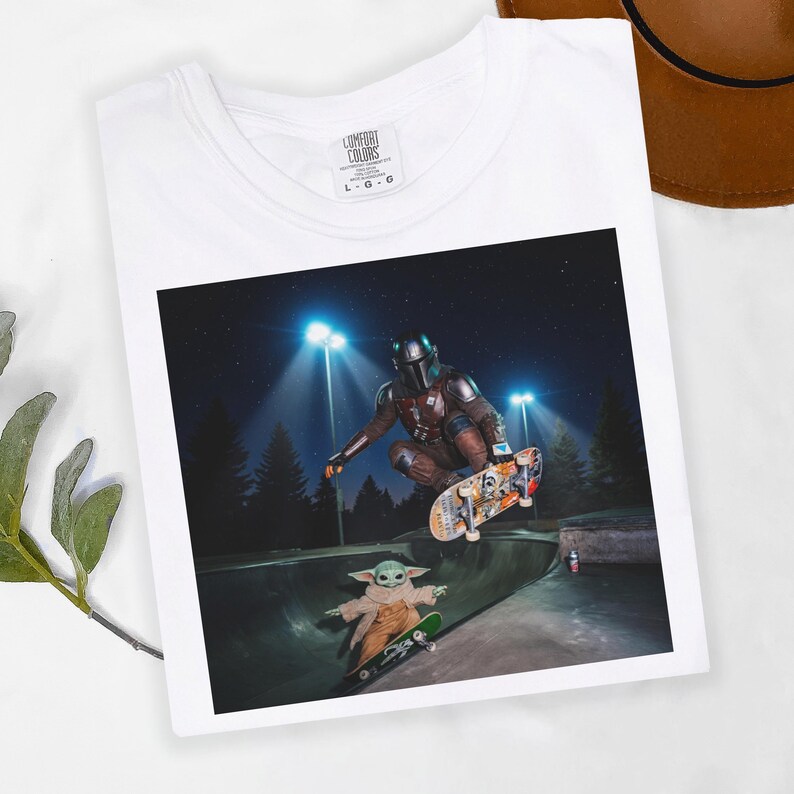 May include: White t-shirt with a graphic of The Mandalorian and Grogu skateboarding in a skate park at night. The shirt has the Comfort Colors logo on the upper left.