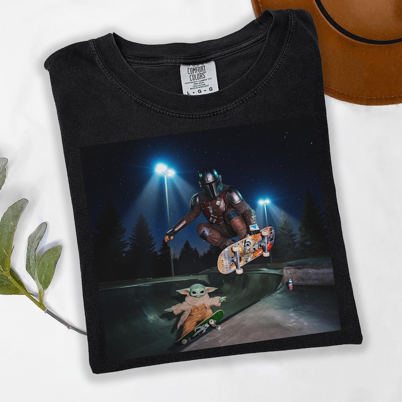 May include: Black Comfort Colors t-shirt featuring a graphic of The Mandalorian and Grogu skateboarding in a skate park at night. The image includes the text "COMFORT COLORS" on the upper right of the shirt.
