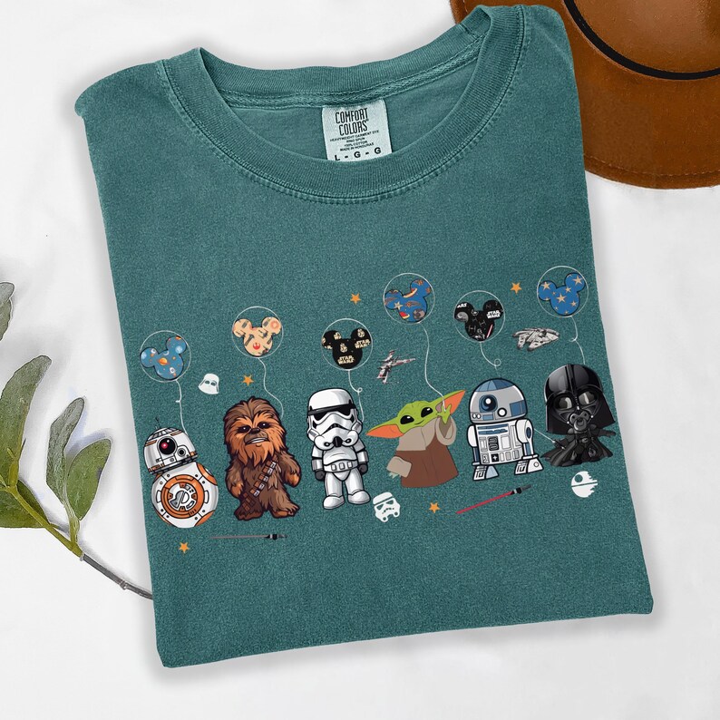 Puede incluir: Camiseta de color verde azulado con personajes de Star Wars y globos tem&aacute;ticos de Mickey Mouse. El dise&ntilde;o muestra a BB-8, Chewbacca, un Stormtrooper, Grogu, R2-D2 y Darth Vader. La etiqueta de la marca Comfort Colors es visible.