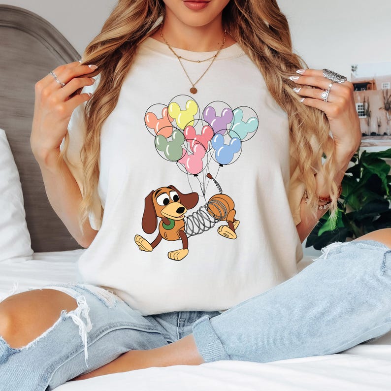 May include: Cream-colored t-shirt featuring a cartoon dachshund with a spring body, holding a bunch of Mickey Mouse-shaped balloons in various pastel colors. The dog is brown, tan, and yellow. The balloons are pink, green, yellow, purple, and blue.