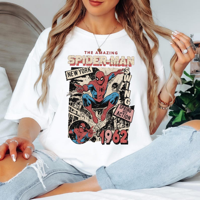 May include: White t-shirt featuring a vintage comic book design of Spider-Man. The graphic includes the text "The Amazing Spider-Man," "New York," and "1962." The superhero is depicted in his classic red and blue suit, mid-swing.