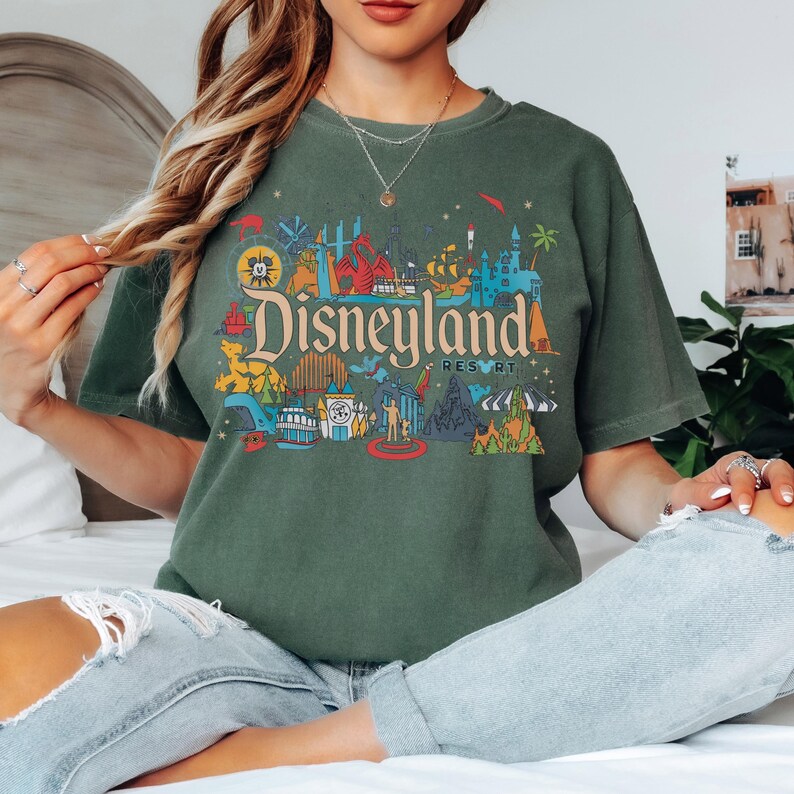 May include: Dark green t-shirt featuring a colorful Disneyland Resort graphic. The design includes iconic park imagery and the Disneyland logo in a vintage style. The shirt is a comfortable, casual style.