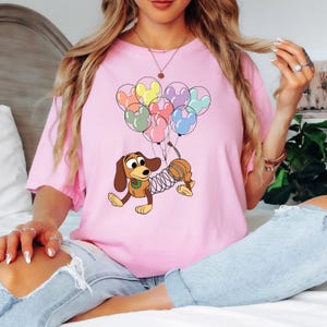May include: Pink t-shirt featuring a cartoon illustration of Slinky Dog from Toy Story. Slinky Dog is holding a bunch of Mickey Mouse-shaped balloons in various colors. The shirt is a light pink color.