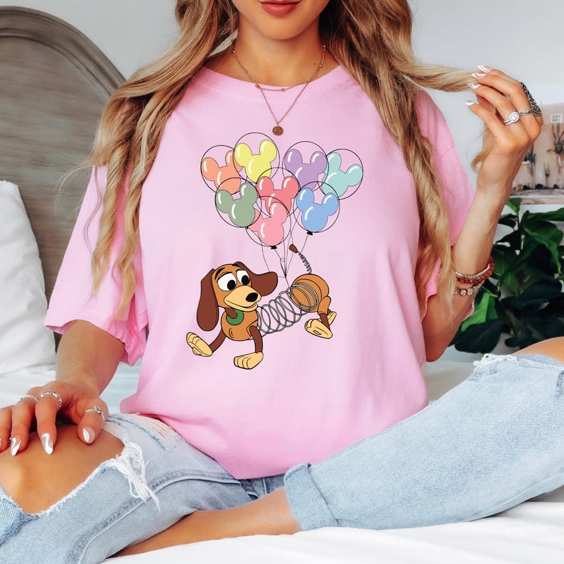 May include: Pink t-shirt featuring a cartoon illustration of Slinky Dog from Toy Story. Slinky Dog is holding a bunch of Mickey Mouse-shaped balloons in various colors. The shirt is a light pink color.