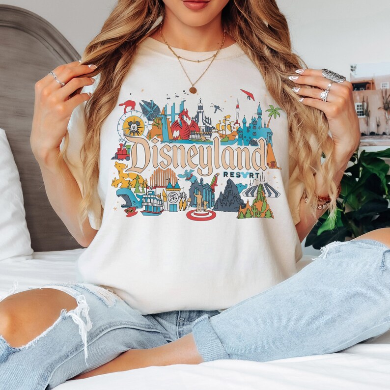 May include: Cream-colored t-shirt featuring a colorful Disneyland Resort graphic. The design includes iconic park imagery and the Disneyland Resort logo in a vintage style. The shirt is a casual, comfortable style.