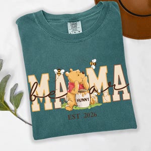 May include: Teal Comfort Colors t-shirt with a graphic of Winnie the Pooh and bees. The shirt says "Mama Bee" with the word "Mama" in large block letters and "bee" in cursive. The shirt also says "EST 2026".