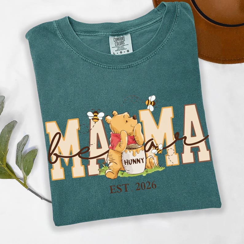 May include: Teal Comfort Colors t-shirt with a graphic of Winnie the Pooh and bees. The shirt says "Mama Bee" with the word "Mama" in large block letters and "bee" in cursive. The shirt also says "EST 2026".