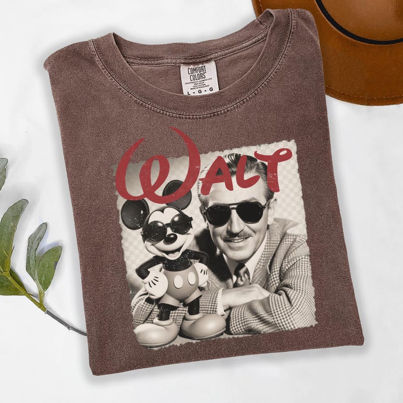 Puede incluir: Una camiseta Comfort Colors marr&oacute;n con un gr&aacute;fico de Walt Disney y Mickey Mouse con gafas de sol. La palabra "Walt" est&aacute; impresa en rojo sobre la imagen. La camiseta est&aacute; doblada.