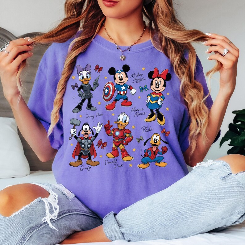 May include: A purple t-shirt featuring a graphic of Disney characters in superhero costumes. Characters include Daisy Duck, Mickey Mouse, Minnie Mouse, Goofy, Donald Duck, and Pluto. The shirt has a relaxed fit.