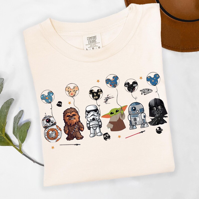 Puede incluir: Camiseta color crema con un gr&aacute;fico de personajes de Star Wars y Disney. El dise&ntilde;o incluye a BB-8, Chewbacca, un Stormtrooper, Grogu, R2-D2 y Darth Vader, cada uno con un globo. La camiseta tiene el logo de Comfort Colors.