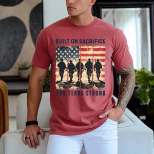 May include: Red t-shirt featuring an American flag design with soldiers walking in silhouette. The text reads "BUILT ON SACRIFICE" above the flag and "250 YEARS STRONG" below. The shirt is worn by a person.