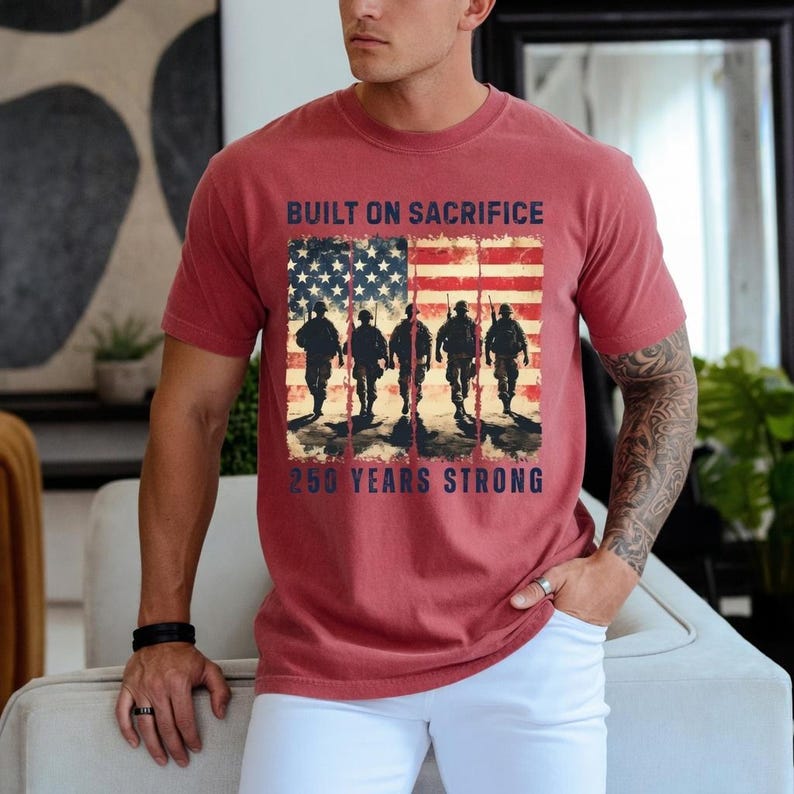 May include: Red t-shirt featuring an American flag design with soldiers walking in silhouette. The text reads "BUILT ON SACRIFICE" above the flag and "250 YEARS STRONG" below. The shirt is worn by a person.
