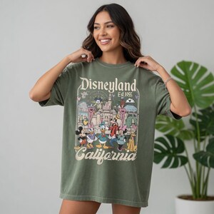 May include: An olive green t-shirt with a Disneyland graphic. The design includes Mickey Mouse, Donald Duck, and other characters, with the text "Disneyland California Est. 1955". The shirt has short sleeves and a relaxed fit. The graphic is colourful.