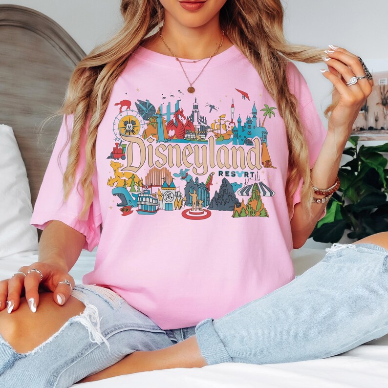 May include: A light pink t-shirt featuring a colorful Disneyland Resort graphic. The graphic includes various park icons and the Disneyland Resort text in a vintage style. The shirt is a short-sleeved crew neck.