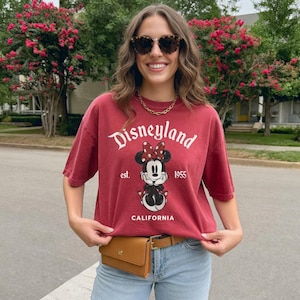 May include: A maroon Disneyland t-shirt featuring Minnie Mouse with a red bow, the text "Disneyland" and "California" in white. The shirt is paired with a brown belt bag and light blue jeans.
