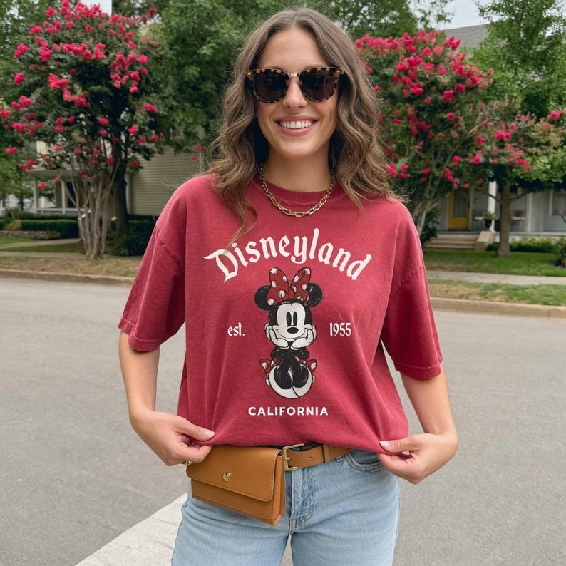 May include: A maroon Disneyland t-shirt featuring Minnie Mouse with a red bow, the text "Disneyland" and "California" in white. The shirt is paired with a brown belt bag and light blue jeans.