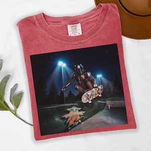 May include: Red Comfort Colors t-shirt featuring a graphic of The Mandalorian and Grogu skateboarding in a skate park at night. The Mandalorian is mid-air on a skateboard, while Grogu rides a smaller board below.