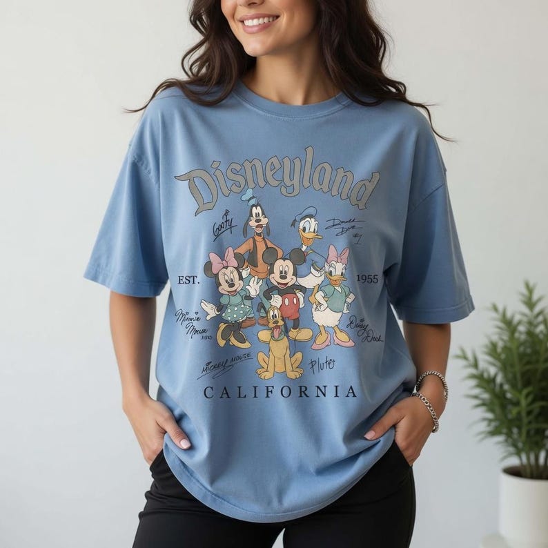 May include: Pale blue t-shirt with the Disneyland logo and Disney characters such as Mickey Mouse, Minnie Mouse, and Goofy. The shirt also features the text "CALIFORNIA" and "EST. 1955".