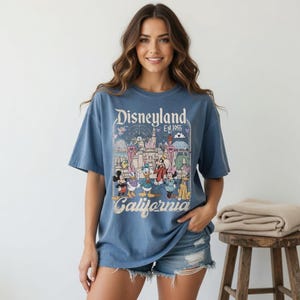 May include: A blue t-shirt with the Disneyland logo and a colourful graphic of Disney characters and park attractions. The shirt includes the text "Disneyland Est. 1955 California".