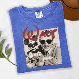 Puede incluir: Una camiseta azul Comfort Colors con un gr&aacute;fico retro de Walt Disney y Mickey Mouse con gafas de sol. La palabra "WALT" est&aacute; impresa en rojo encima de la imagen.