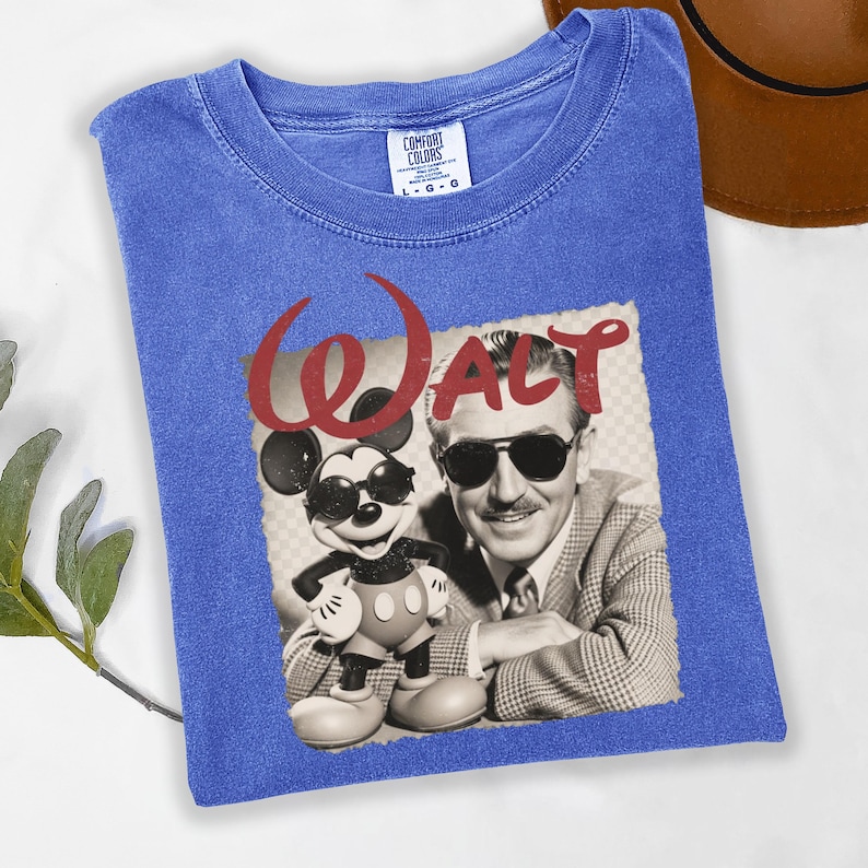 Puede incluir: Una camiseta azul Comfort Colors con un gr&aacute;fico retro de Walt Disney y Mickey Mouse con gafas de sol. La palabra "WALT" est&aacute; impresa en rojo encima de la imagen.