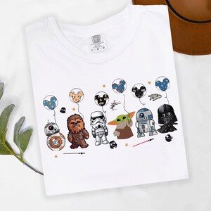 Puede incluir: Camiseta blanca con personajes de dibujos animados de Star Wars y Disney. El dise&ntilde;o incluye a BB-8, Chewbacca, un Stormtrooper, Grogu, R2-D2 y Darth Vader, cada uno sosteniendo un globo. Los globos tienen dise&ntilde;os de Mickey Mouse y Star Wars.