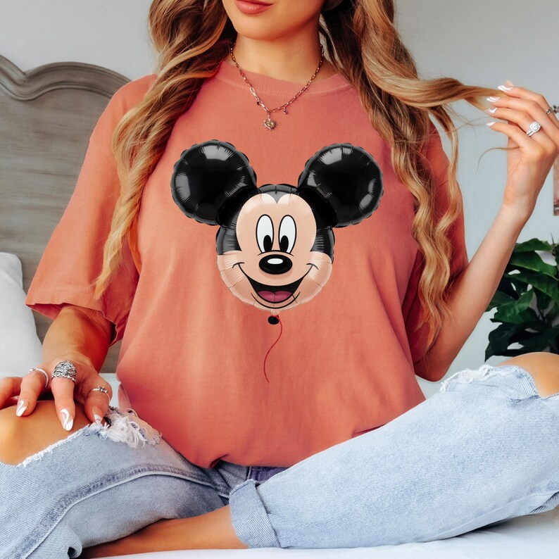 Puede incluir: Camiseta de color coral con un dise&ntilde;o de globo de Mickey Mouse. El globo tiene orejas negras y una cara sonriente con una cuerda roja. La camiseta es de estilo casual.