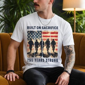 May include: White t-shirt featuring a graphic of the American flag with silhouettes of soldiers. The text "BUILT ON SACRIFICE" is above the flag, and "250 YEARS STRONG" is below. The shirt is worn by a person.
