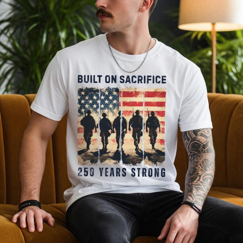 May include: White t-shirt featuring a graphic of the American flag with silhouettes of soldiers. The text "BUILT ON SACRIFICE" is above the flag, and "250 YEARS STRONG" is below. The shirt is worn by a person.