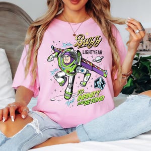 Puede incluir: Camiseta rosa claro con un dise&ntilde;o de Buzz Lightyear. El dise&ntilde;o muestra a Buzz con su traje espacial, con las palabras "Buzz Lightyear" y "To Infinity and Beyond".