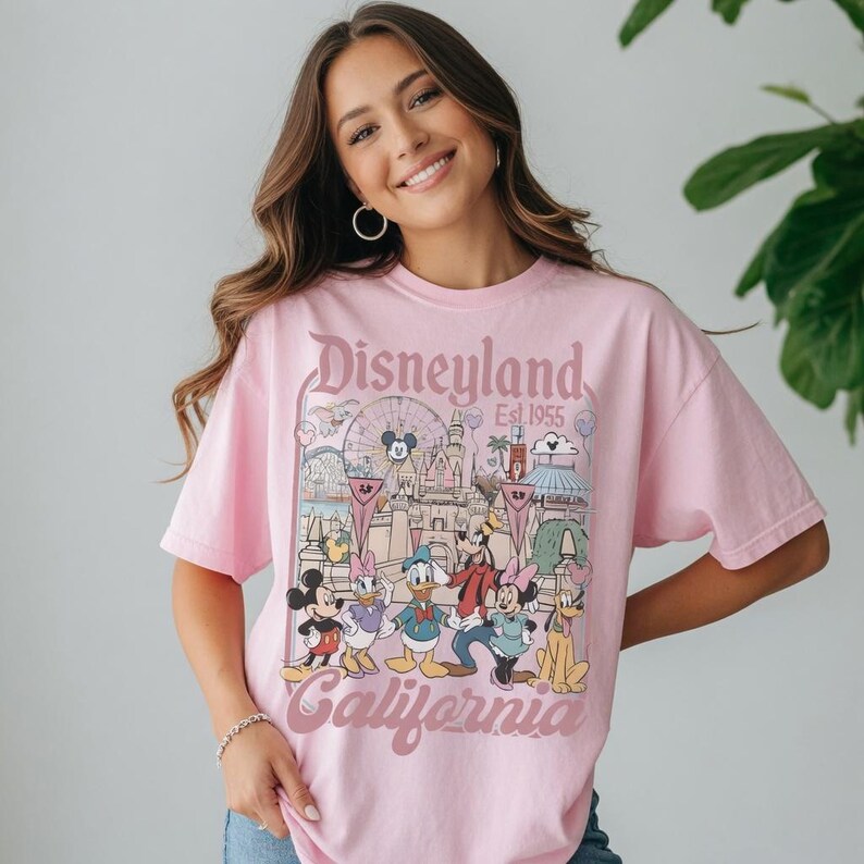 May include: A light pink t-shirt with a Disneyland graphic. The design includes Mickey Mouse, Donald Duck, and other characters. The text "Disneyland Est. 1955 California" is printed on the front.