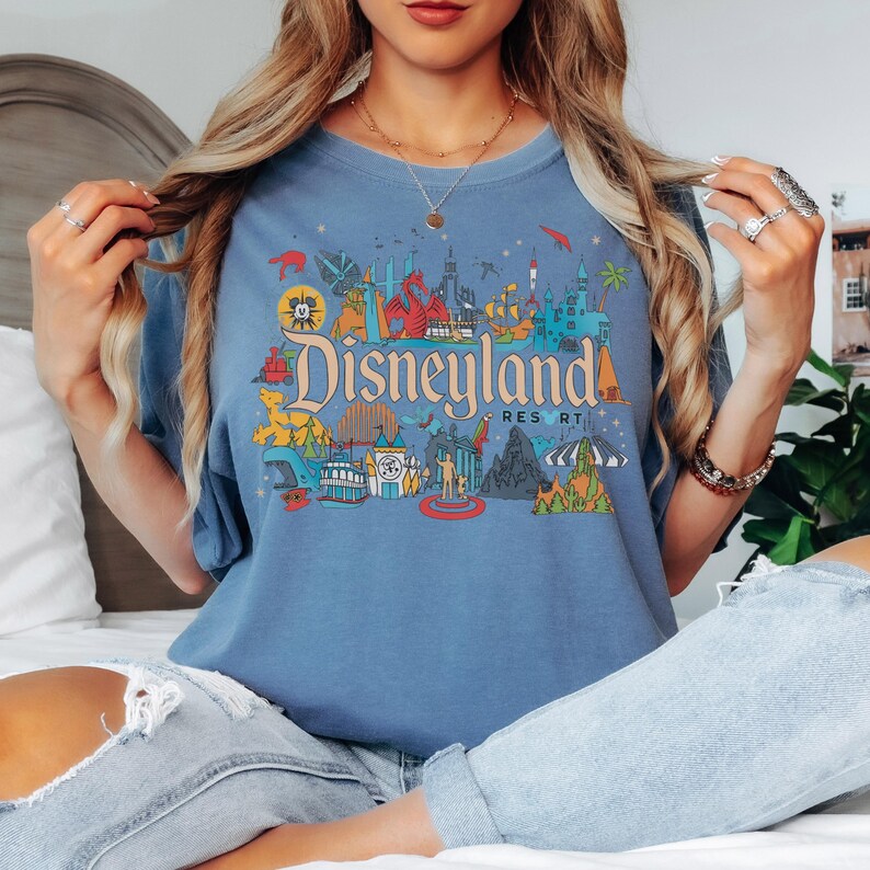 May include: A blue t-shirt featuring a colorful Disneyland Resort graphic. The design includes iconic park imagery and the Disneyland logo in orange and teal. The shirt is a comfortable, casual style.