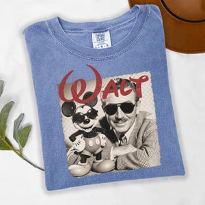 Puede incluir: Camiseta azul con un gr&aacute;fico de Walt Disney y Mickey Mouse con gafas de sol. La palabra "WALT" est&aacute; impresa en rojo encima de la imagen. La camiseta es de un material suave.