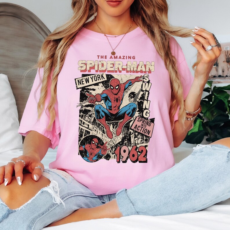 May include: Pink t-shirt featuring a vintage comic book graphic of Spider-Man. The design includes the text "THE AMAZING SPIDER-MAN NEW YORK 1962" and the phrase "SWING INTO ACTION". The graphic is in red, blue, and black.