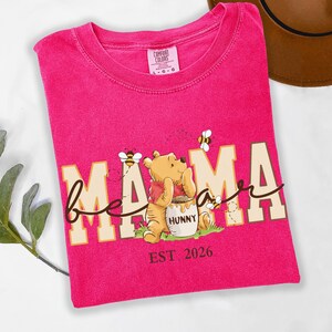 May include: A hot pink t-shirt with the words "bee mama" in a whimsical font, featuring a cartoon bear and bees. The shirt also has the text "EST 2026". The shirt is made of a soft material.