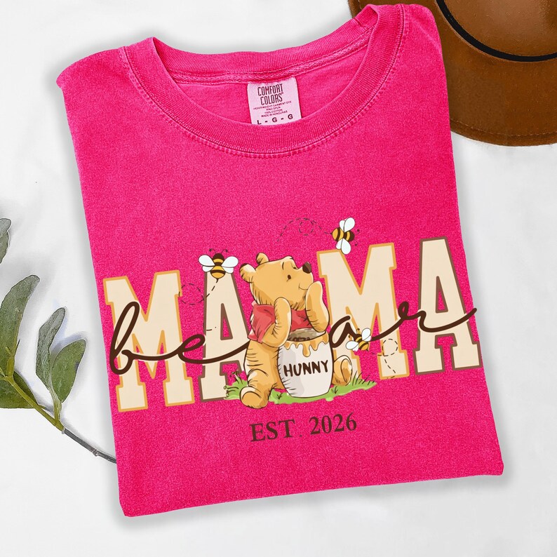 May include: A hot pink t-shirt with the words "bee mama" in a whimsical font, featuring a cartoon bear and bees. The shirt also has the text "EST 2026". The shirt is made of a soft material.