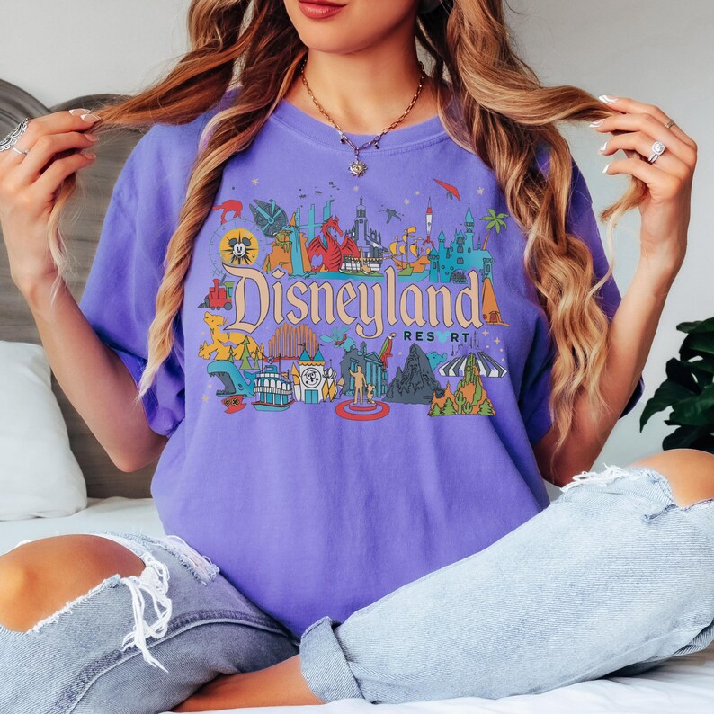 May include: A purple t-shirt featuring a colorful Disneyland Resort graphic. The design includes iconic park imagery and the Disneyland logo in a vintage style. The shirt is a short-sleeved, crew neck style.