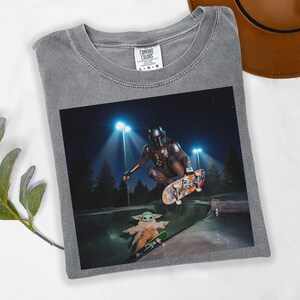 May include: Gray Comfort Colors t-shirt featuring a graphic of The Mandalorian and Grogu skateboarding in a skate park at night. The image shows the characters mid-air on skateboards, with bright lights and trees in the background.