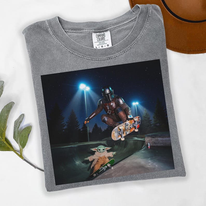 May include: Gray Comfort Colors t-shirt featuring a graphic of The Mandalorian and Grogu skateboarding in a skate park at night. The image shows the characters mid-air on skateboards, with bright lights and trees in the background.