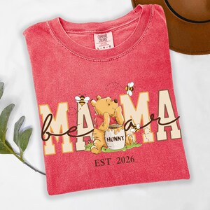 May include: Red t-shirt with the words "bee mama bear" and a cartoon illustration of Winnie the Pooh with a honey pot and bees. The text "EST 2026" is also printed on the shirt. The shirt is made by Comfort Colors.