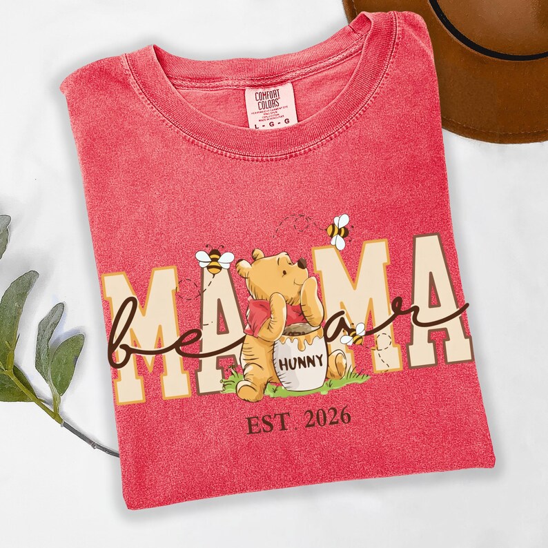May include: Red t-shirt with the words "bee mama bear" and a cartoon illustration of Winnie the Pooh with a honey pot and bees. The text "EST 2026" is also printed on the shirt. The shirt is made by Comfort Colors.
