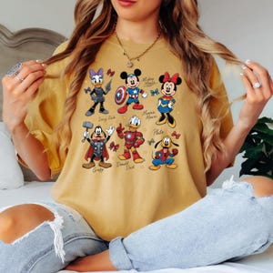 May include: Mustard yellow t-shirt featuring cartoon characters Daisy Duck, Mickey Mouse, Minnie Mouse, Goofy, Donald Duck, and Pluto, each dressed as superheroes. The shirt has a crew neck and short sleeves. The characters are in various action poses.