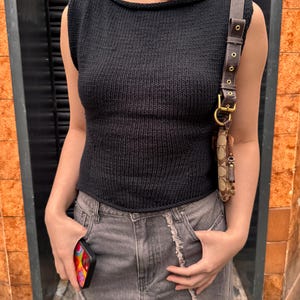 Peut inclure: Un haut noir sans manches en tricot avec un col bateau. Le haut est associé à une jupe en jean grise. Un sac à bandoulière en cuir marron avec une boucle dorée et une coque de téléphone avec un motif coloré sont également visibles.