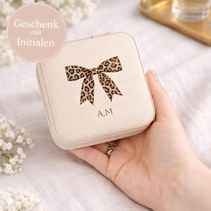 Könnte beinhalten: Eine beige quadratische Schmuckschatulle mit einem Leopardenmuster-Schleifendesign und den Initialen "A.M.". Der Text "Geschenk mit Initialen" befindet sich in einem rosa Kreis. Die Box wird in einer Hand gehalten.