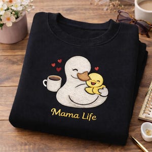 May include: Black sweatshirt with an embroidered design featuring a white duck embracing a yellow duckling. A coffee cup and red hearts are also embroidered. The words "Mama Life" are embroidered in gold.