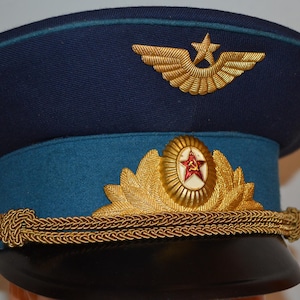 Op de afbeelding: Een donkerblauwe militaire pet met een zwarte klep, een lichtblauwe band en een goudkleurig gevlochten koord. De pet is voorzien van een gouden embleem met een gevleugelde ster op de bovenkant en een gouden lauwerkrans met een rode ster.