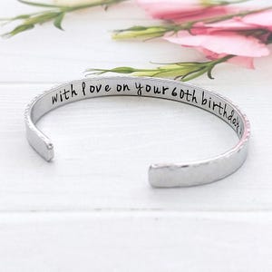 May include: A silver cuff bracelet engraved with the words "with love on your 60th birthday". The open-ended bracelet has a textured finish. Pink flowers are visible in the background.